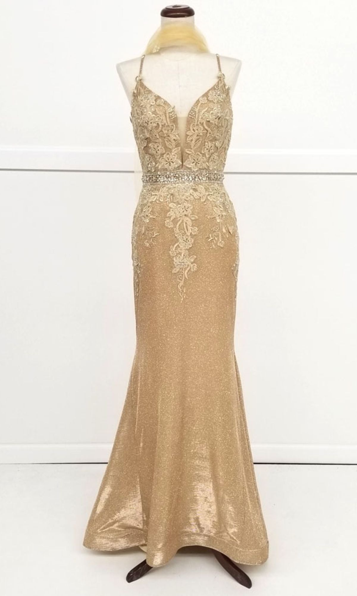 The Chicas C243 is a gold, floor-length mermaid prom dress with a fitted silhouette, floral lace appliqué bodice, thin straps, embellished waistband, and subtle shimmer, shown on a dress form.